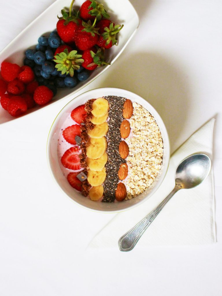 A breakfast bowl with ingredients arranged in rows, including strawberries, bananas, chia, almonds, and oats. Another bowl of berries is to the side.