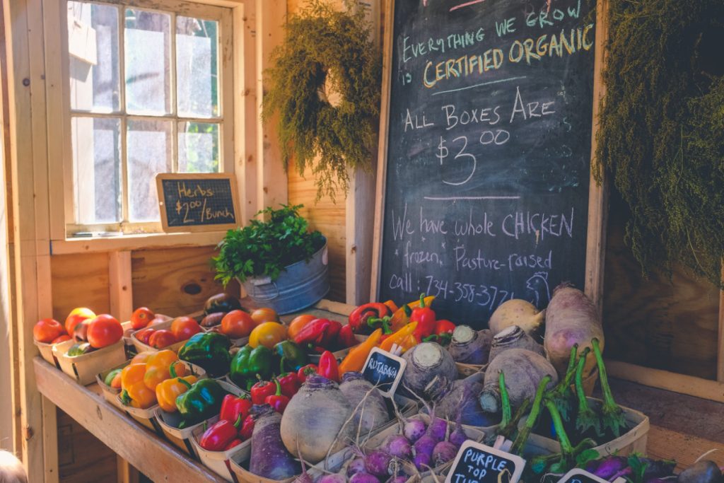 An organic farm stand with a blackboard. The farm stand is selling yellow, green, and red peppers, rutabaga, eggplants, and herbs.